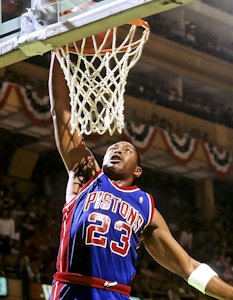 رجل يرتدي قميصًا أزرق اللون مكتوب عليه كلمة "PISTONS" والرقم "23" في الأمام، يحاول غمس كرة السلة.