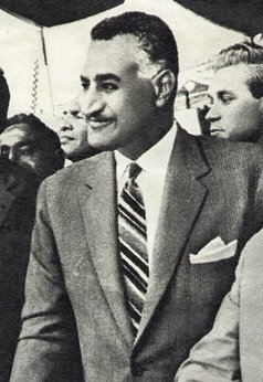 رجل مبتسم ذو شارب أسود يواجه يسار المشاهد. شعره داكن وقصير، أبيض عند الصدغين. يرتدي بدلة رمادية من قطعتين على الطراز الغربي وقميصًا أبيض، مع ربطة عنق مخططة بزوايا حادة ومنديل جيب أبيض ظاهر. خلفه تظهر عدة وجوه.
