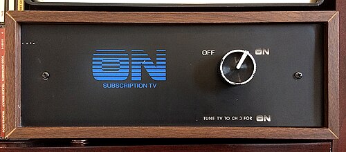 صورة أمامية لصندوق خشبي ذي واجهة سوداء مغطاة بقشرة خشبية. يزين الواجهة شعار "ON Subscription TV" باللون الأزرق، حيث تظهر حروف "ON" بتدرج لوني خطي. يوجد زر فضي للاختيار بين خيارين: إيقاف التشغيل (OFF) وتشغيل (ON) (بتصميم مشابه للشعار). أسفل الزر توجد عبارة "اضبط التلفزيون على القناة 3 للتشغيل".