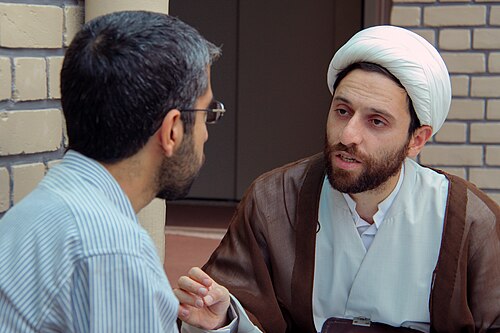 رجل يرتدي عمامة وملابس دينية فضفاضة يتحدث مع رجل يرتدي قميصاً بأزرار ويستمع إليه باهتمام.