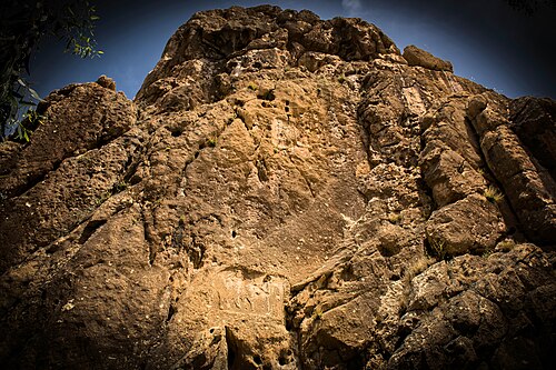 The relief is located on the top of a cliff towering over the village of Sarpol-e Zahab. A second relief from the Parthian Empire period appears below.