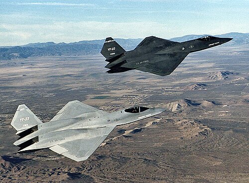 نموذجان إنتاجيان من طراز YF-23 يحلقان فوق الصحراء، الأول يُدعى "الشبح الرمادي" ومطلي باللون الرمادي الفحمي ويقع على يسار الثاني المسمى "العنكبوت" والمطلي باللون الرمادي الفاتح، والذي يقع في المقدمة مباشرة.