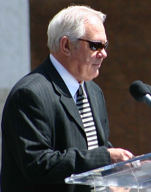 رود فيليبس يرتدي نظارة شمسية سوداء ويتحدث إلى حشد من الناس على منصة مزودة بميكروفون