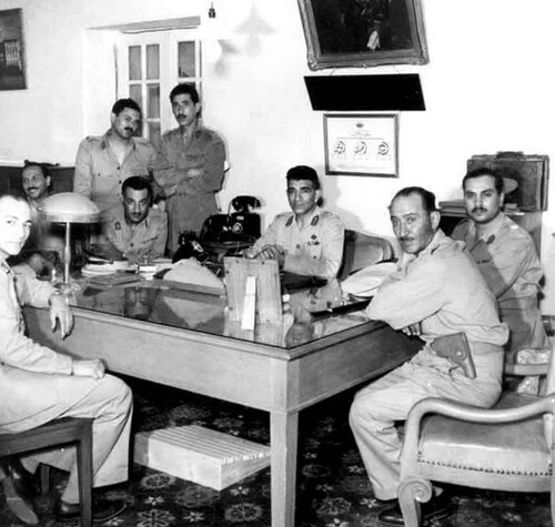 ثمانية رجال يرتدون الزي العسكري، يقفون في غرفة حول طاولة مستطيلة. جميع الرجال جالسون باستثناء الشخصين الثالث والخامس من اليسار. أما الشخصان الثالث والخامس من اليسار فهما واقفان.