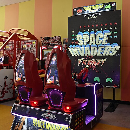 كرسيان أحمران مثبتان على قاعدة يواجهان شاشة كبيرة تعرض لعبة "Space Invaders Frenzy". وُضعت مدافع بلاستيكية مضيئة أمام الكرسيين موجهة نحو الشاشة.