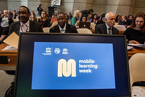 تعرض شاشة كمبيوتر في مقدمة غرفة تضم صناع السياسات شعار أسبوع التعلم عبر الهاتف المحمول