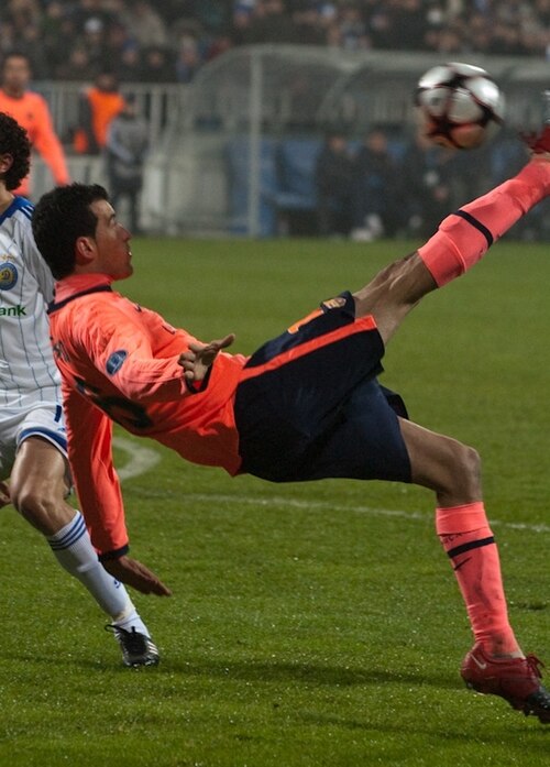 لاعب في الهواء. ينظر إلى الكرة؛ ساقه تتحرك لأعلى باتجاه الكرة، ويبدو كما لو أنها ستضرب الكرة فوق رأس اللاعب وخلفه.