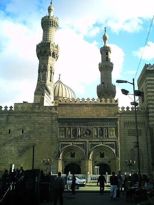 يطل مدخلان مقوسان في رواق مبنى كبير من طابقين على شارع. وفوق القوسين، يزدان جدار المبنى بنقوش وزخارف. وإلى اليمين، يرتفع المبنى إلى طابق ثالث. وخلف الجدار، تظهر مئذنتان تُحيطان بقمة قبة.