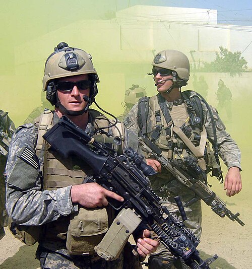 مايكل منصور، أحد أفراد قوات البحرية الأمريكية الخاصة (SEAL)، برفقة زميل له من نفس الفريق، يرتديان زيًا مموهًا أخضر اللون، بالإضافة إلى بزات قتالية خضراء. يحمل كلاهما أسلحة نارية ويرتديان نظارات شمسية. يظهر في الخلفية مبنى أبيض اللون ودخان أخضر يتصاعد.