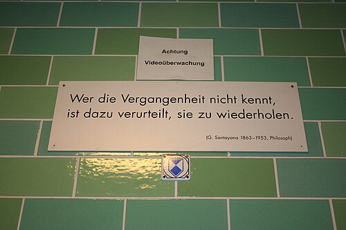 جدار من الطوب الأخضر عليه لافتة بيضاء مكتوب عليها "Wer die Vergangenheit nicht kennt, / ist dazu verurteilt, sie zu wiederholden. / (ج. سانتايانا 1863–1953، فيلسوف)