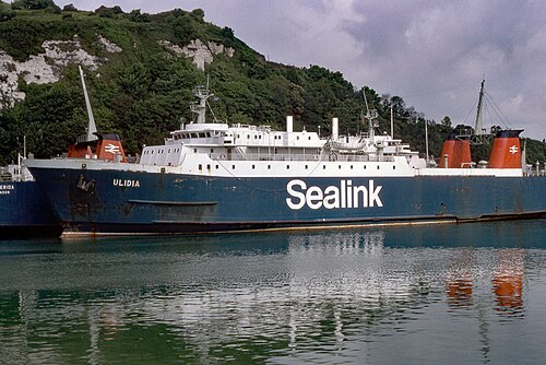 عبّارة راسية، تُرى من الجهة المقابلة للماء. هيكلها مطلي باللون الأزرق، وعليه كلمة "Sealink" بخط serif. في الخلفية، تظهر منحدرات طباشيرية مغطاة بنباتات كثيفة.