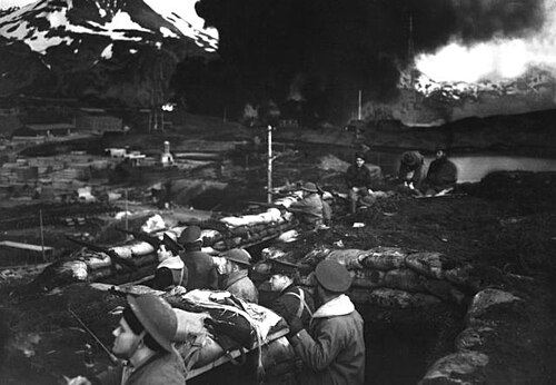 alt=صورة تاريخية لجنود مشاة البحرية الأمريكية في خنادق دفاعية خلال الهجمات اليابانية عام 1942، بينما تحترق خزانات الوقود في الخلفية.
