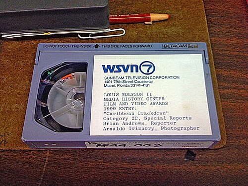 A videocassette on top of a clipboard next to its plastic case, a grease pencil and a paper clip. The label on the cassette contains the WSVN letterhead: "WSVN 7 Sunbeam Television Corporation, 1401 79th Street Causeway, Miami, Florida 33141-4181" then in print below, "LOUIS WOLFSON II MEDIA HISTORY CENTER FILM AND VIDEO AWARDS 1989 ENTRY: 'Caribbean Crackdown' Category 2C, Special Reports, Brian Andrews, Reporter, Arnaldo Irizarry, Photgrapher"