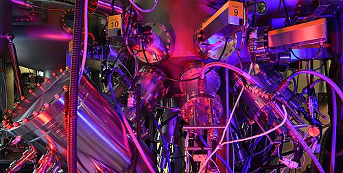 Close-up of a complex stainless-steel scientific instrument with multiple vacuum chambers, circular flanges, cables, and labeled ports, illuminated by vivid purple and red lighting, suggesting a high-precision laboratory system such as molecular beam epitaxy or vacuum deposition equipment.