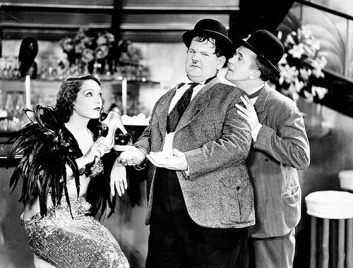 لقطة ثابتة من فيلم أبيض وأسود تظهر لوبي فيليز جالسة وترتدي ريشًا مع لوريل وهاردي في مشهد من فيلم حفلة هوليوود (1934).