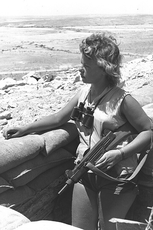 امرأة إسرائيلية مسلحة ببندقية عوزي تقوم بواجب الحراسة في النقب (1956)، لاحظ المقبض الخشبي