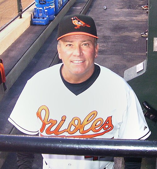 رجل يرتدي قميص بيسبول أبيض مكتوب عليه "Orioles" بخط برتقالي على الصدر، وقبعة بيسبول سوداء عليها صورة طائر الأوريول البرتقالي في الأمام.