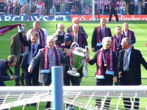 في المقدمة رجلان يحملان كأسًا كبيرًا، يرتديان أوشحة بلون النبيذ الأحمر وميدالية حول أعناقهما. ويحيط بهما عشرة لاعبين قدامى يرتدون بدلات رسمية وميداليات وأوشحة حول أعناقهم.