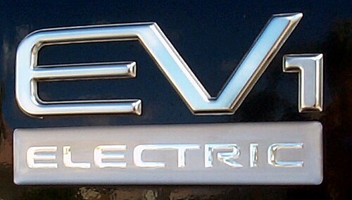 شعار يحمل النص "EV1" بأحرف كبيرة معدنية ومنمقة. أسفل ذلك، توجد كلمة "ELECTRIC" بأحرف بيضاء صغيرة وكبيرة.