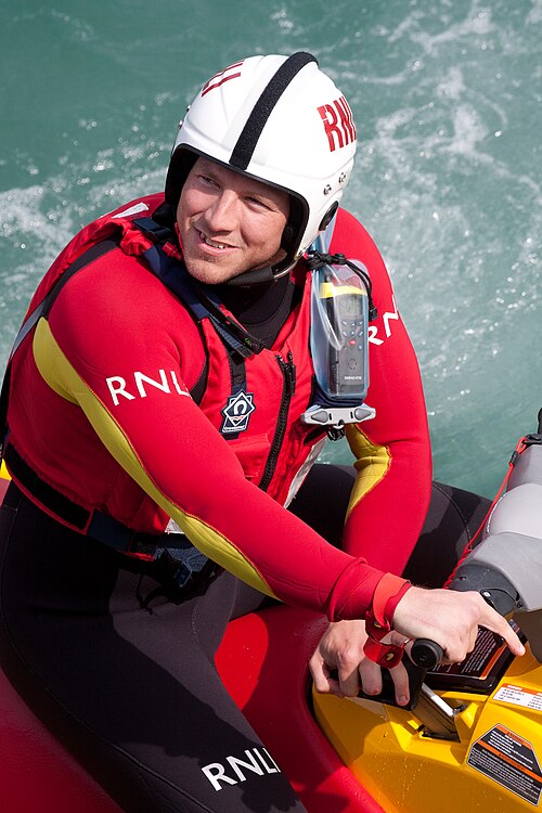 أحد أفراد طاقم RNLI يجلس في قارب مطاطي صغير، ويرتدي خوذة وبدلة غطس حمراء زاهية.