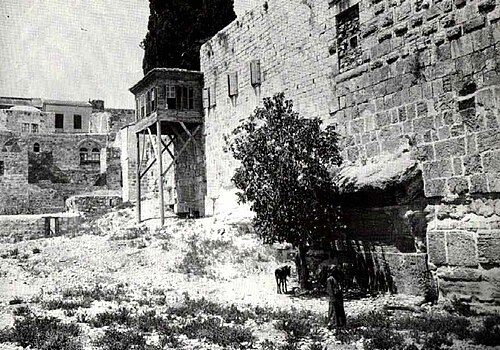 صورة فوتوغرافية بالأبيض والأسود تُظهر رقعة أرض وعرة مع جدار حجري يمتد على طول الجانب الأيمن وسلسلة من الأحجار البارزة في أسفل الجدار في المقدمة، محجوبة جزئيًا بشجيرة