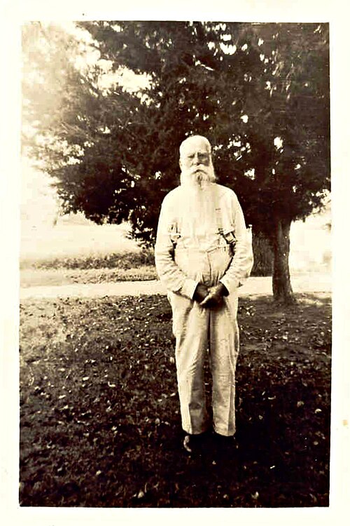 صورة بالأبيض والأسود (حوالي عام 1900) لرجل مسن ذي شعر أبيض ولحية طويلة يرتدي قميصاً بأكمام طويلة وحمالات، يقف أمام شجرة.