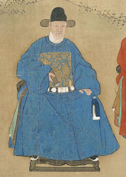 لي دونغيانغ جالس على كرسي، يرتدي رداءً رسمياً أزرق اللون مع شارة رتبة مطرزة وقبعة رسمية سوداء.